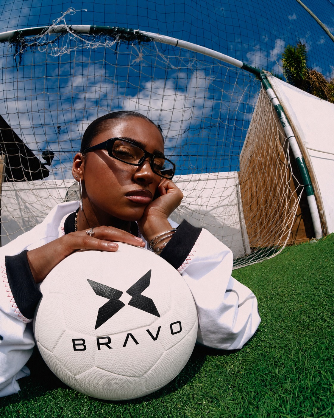 Woman resting on a BRAVO soccer ball on an outdoor pitch under a blue sky